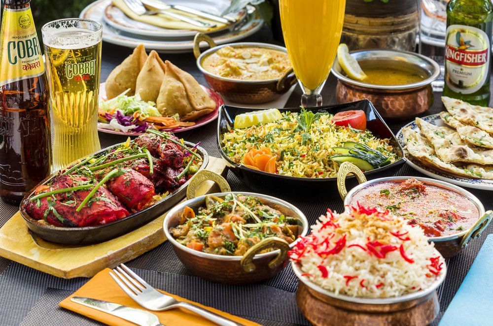 Se sirve comida india en una mesa: diversos curris, arroz, samosas y naan, con bebidas como cerveza y un lassi de mango.