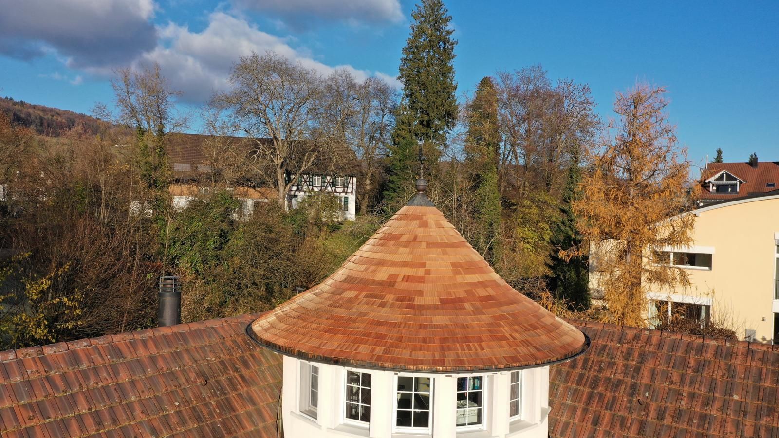 Draufsicht auf einen weissen, runden Turm mit einem rotbraunen, kegelförmigen Dach, umgeben von Bäumen und Gebäuden.