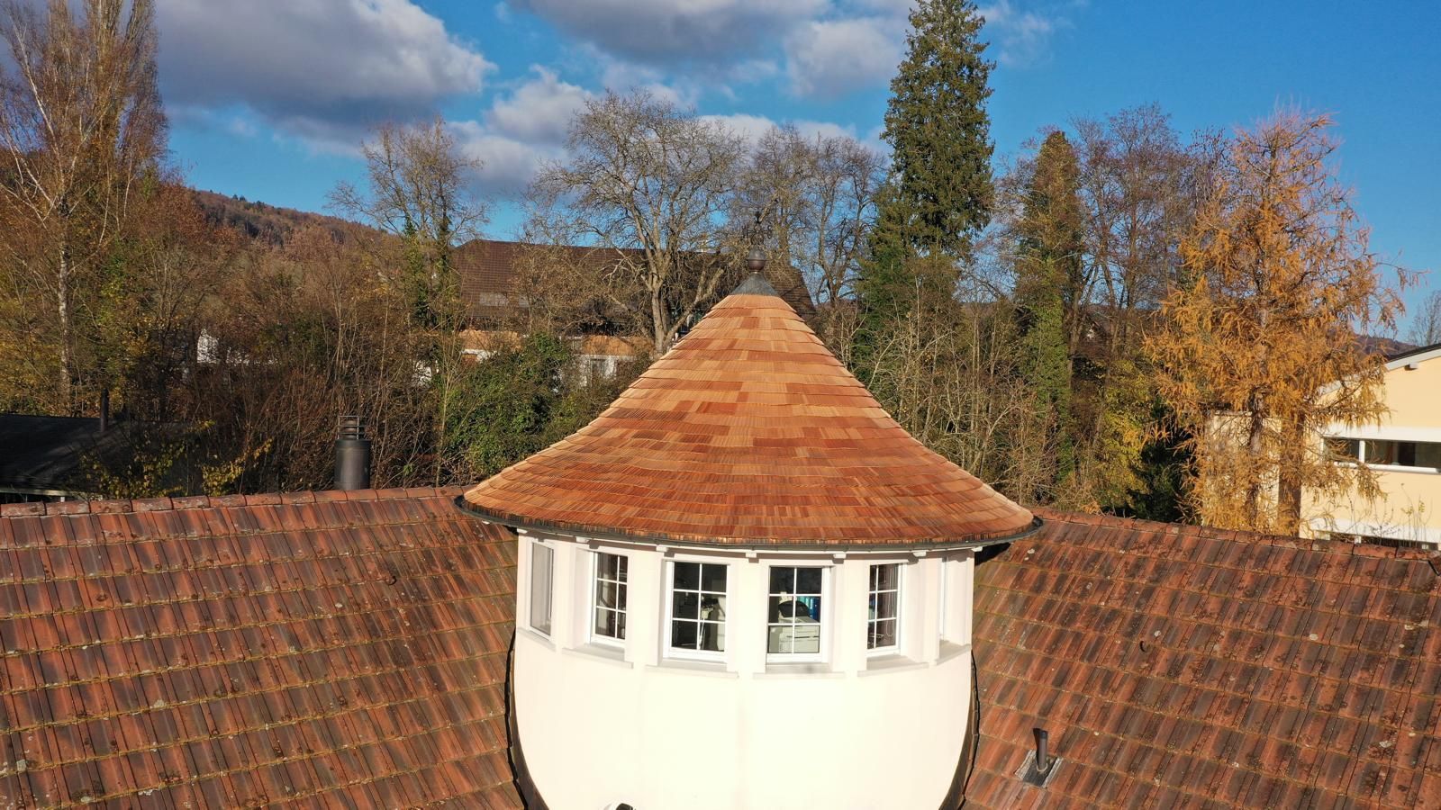 Weisser Turm mit rotem Ziegeldach und geschwungenem Dach. Herbstbäume im Hintergrund.