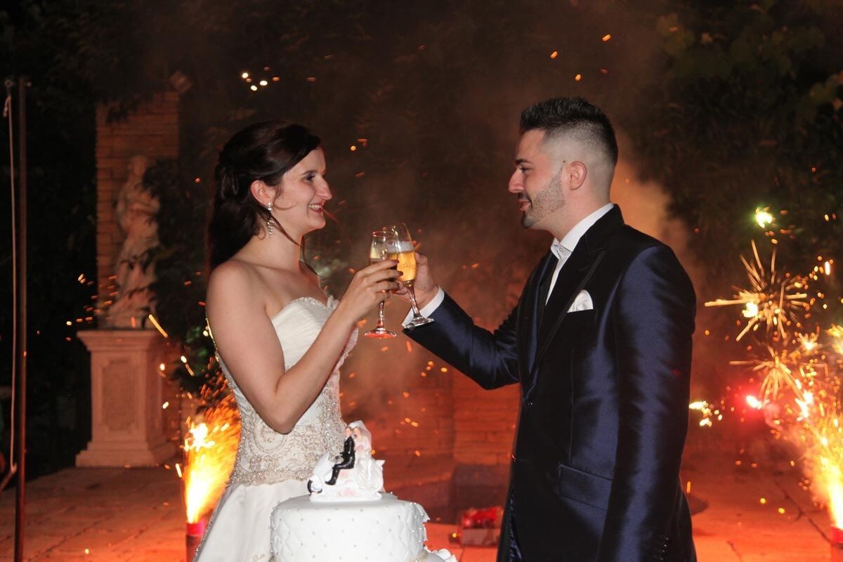 Casal brindando com champanhe na recepção de casamento com fogos de artifício.