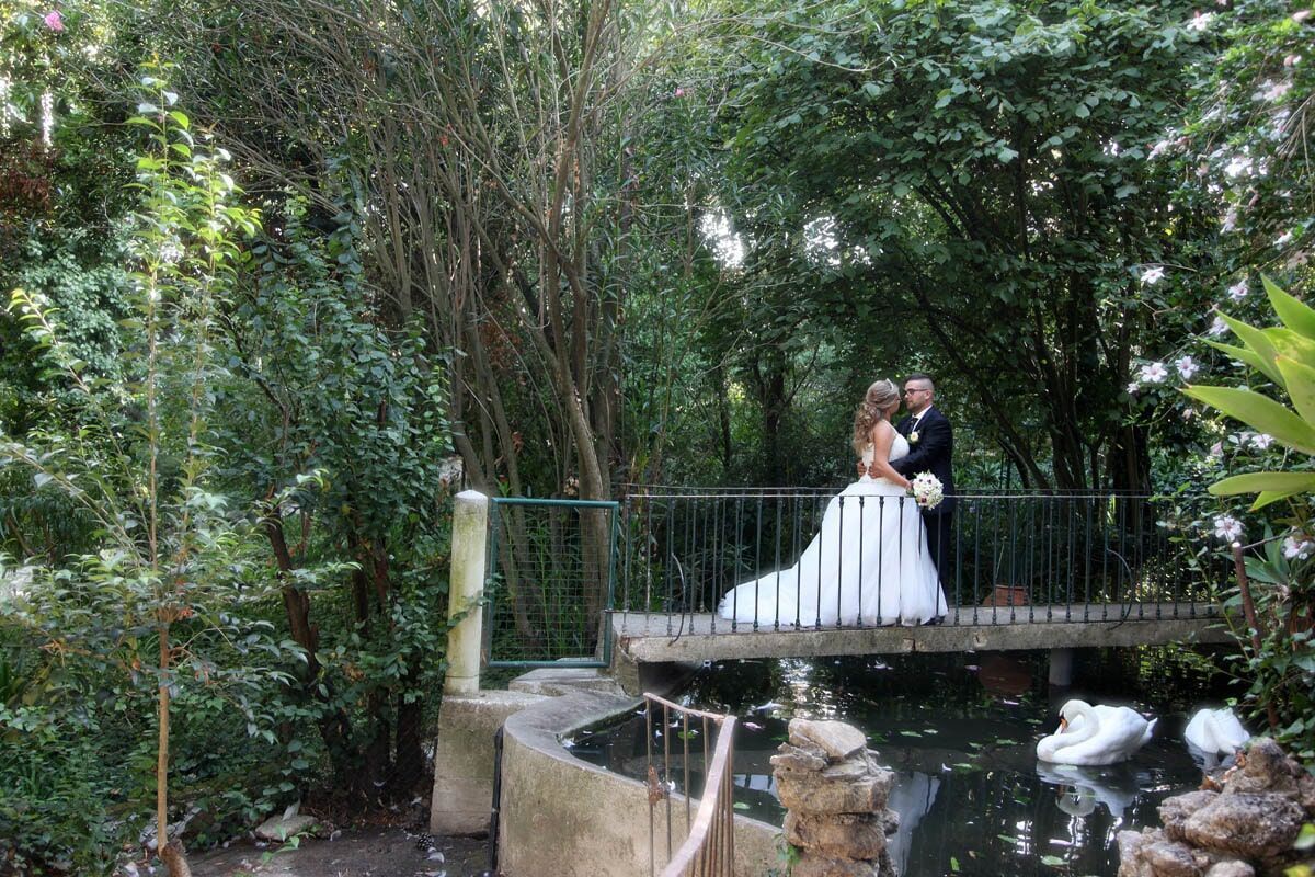 Os noivos posam em uma ponte sobre um lago, rodeados por uma vegetação exuberante.