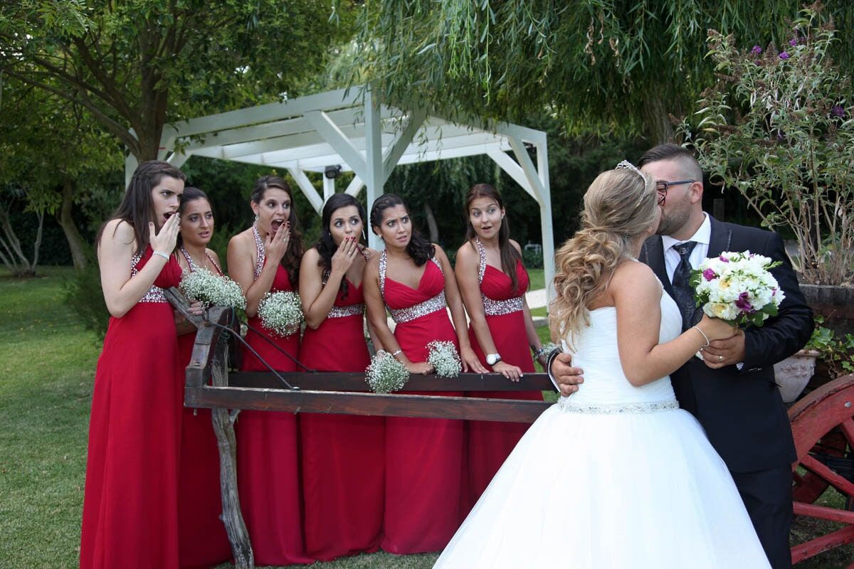 Noivos se beijam ao ar livre; damas de honra de vestido vermelho reagem com surpresa.