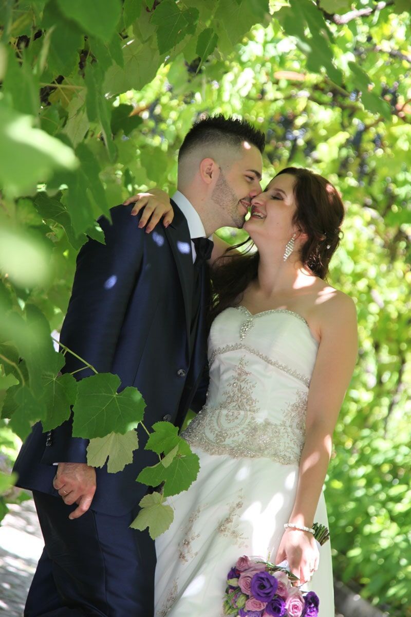 Noivos se beijando sob folhagem verde; o noivo veste um terno azul-marinho, a noiva um vestido branco, segurando um buquê.