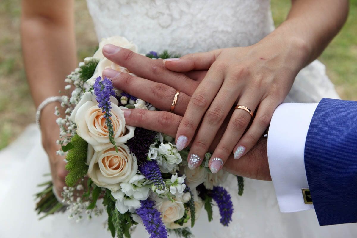As mãos da noiva e do noivo entrelaçadas sobre um buquê, exibindo as alianças de casamento.