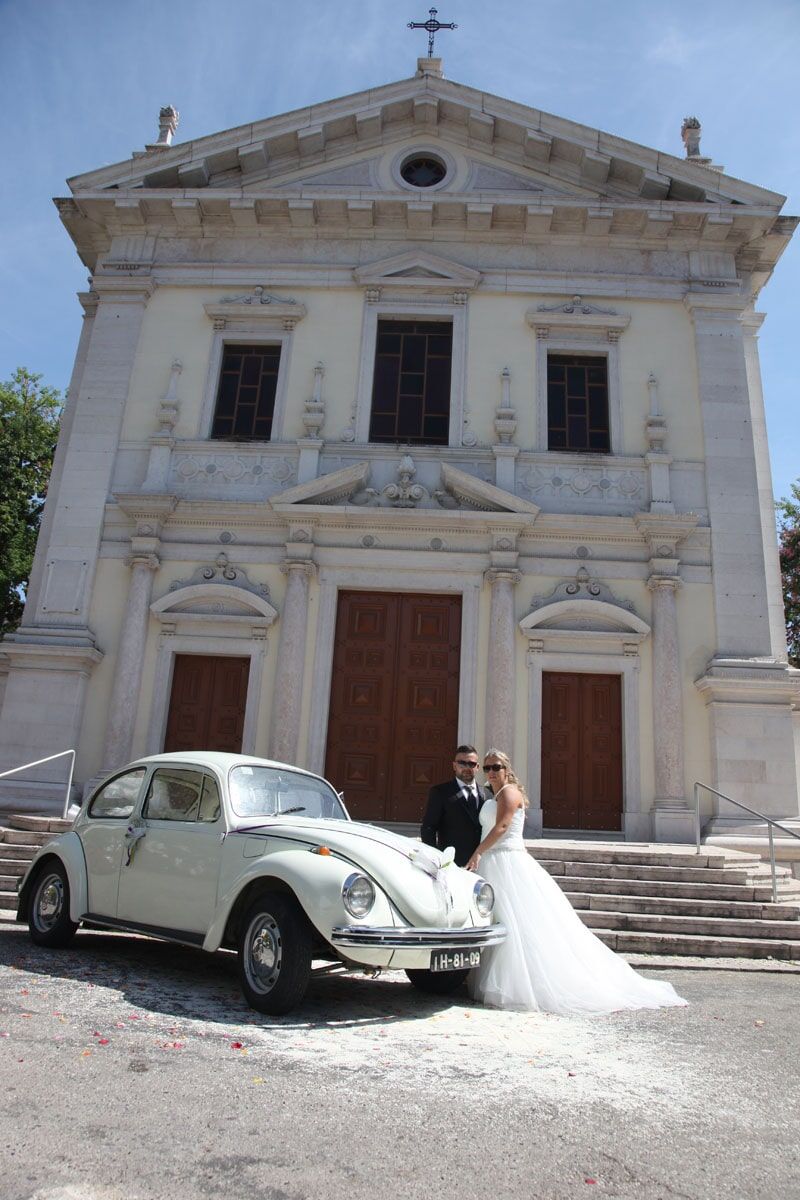 Os recém-casados posam ao lado de um carro clássico branco em frente a uma igreja. A noiva veste um vestido branco.