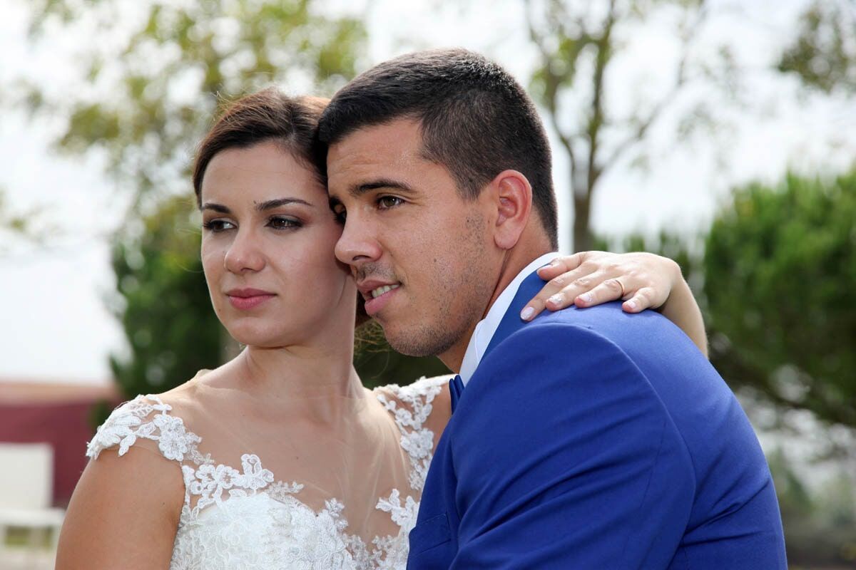 Noivos se abraçam ao ar livre; o noivo de terno azul, a noiva de vestido branco com detalhes em renda.