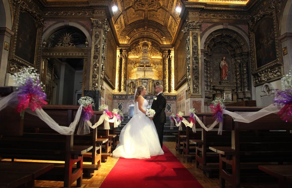 Os noivos estão em uma igreja decorada, com tapete vermelho no corredor, altar ornamentado e bancos decorados com flores.
