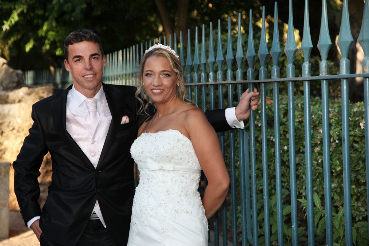Noivos posando junto a uma cerca de ferro forjado; a noiva com um vestido branco sem alças e o noivo com um terno preto.