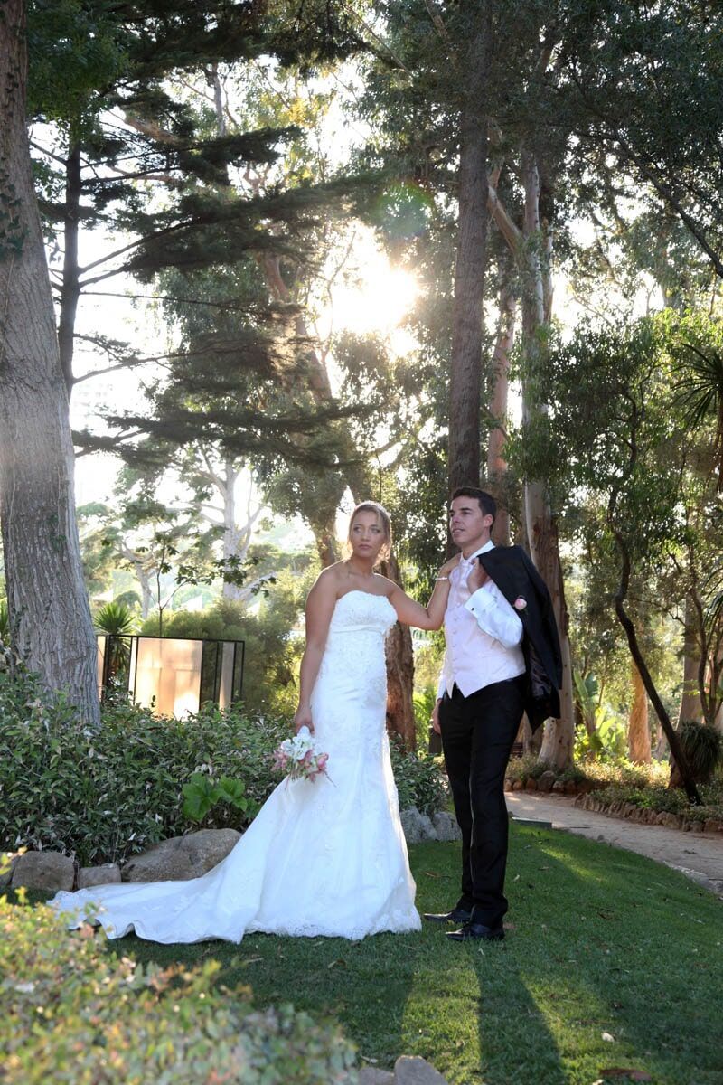 Noivos posam ao ar livre sob a luz do sol, a mulher com um vestido branco e o homem com um paletó, cercados por árvores.