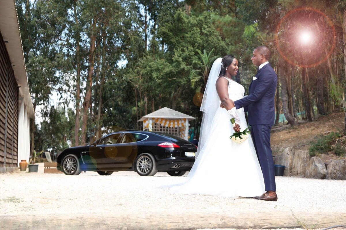 Casal em trajes de casamento se abraça, com um carro preto estacionado próximo, ao ar livre.