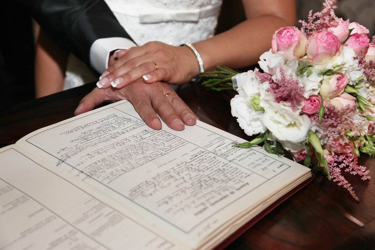 Mãos entrelaçadas sobre um documento aberto, ao lado de um buquê de flores.