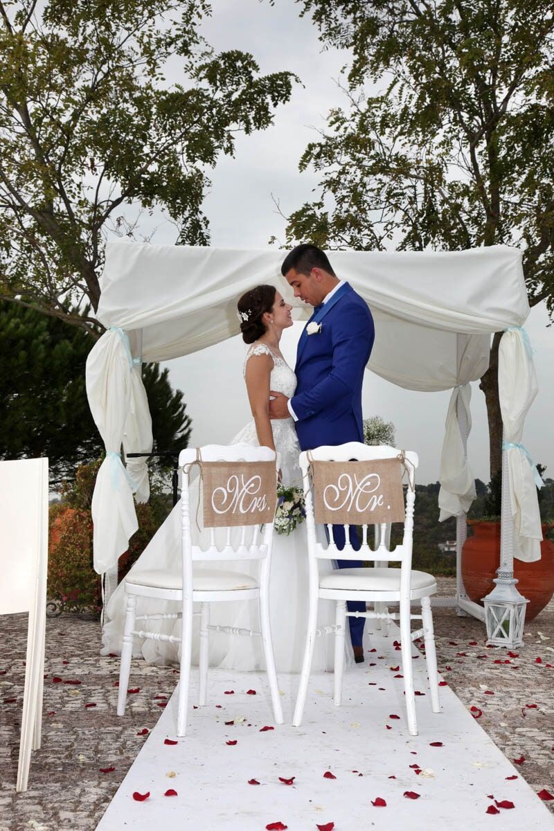 Casal em cerimônia de casamento sob um dossel branco. Noiva de vestido branco, noivo de terno azul.