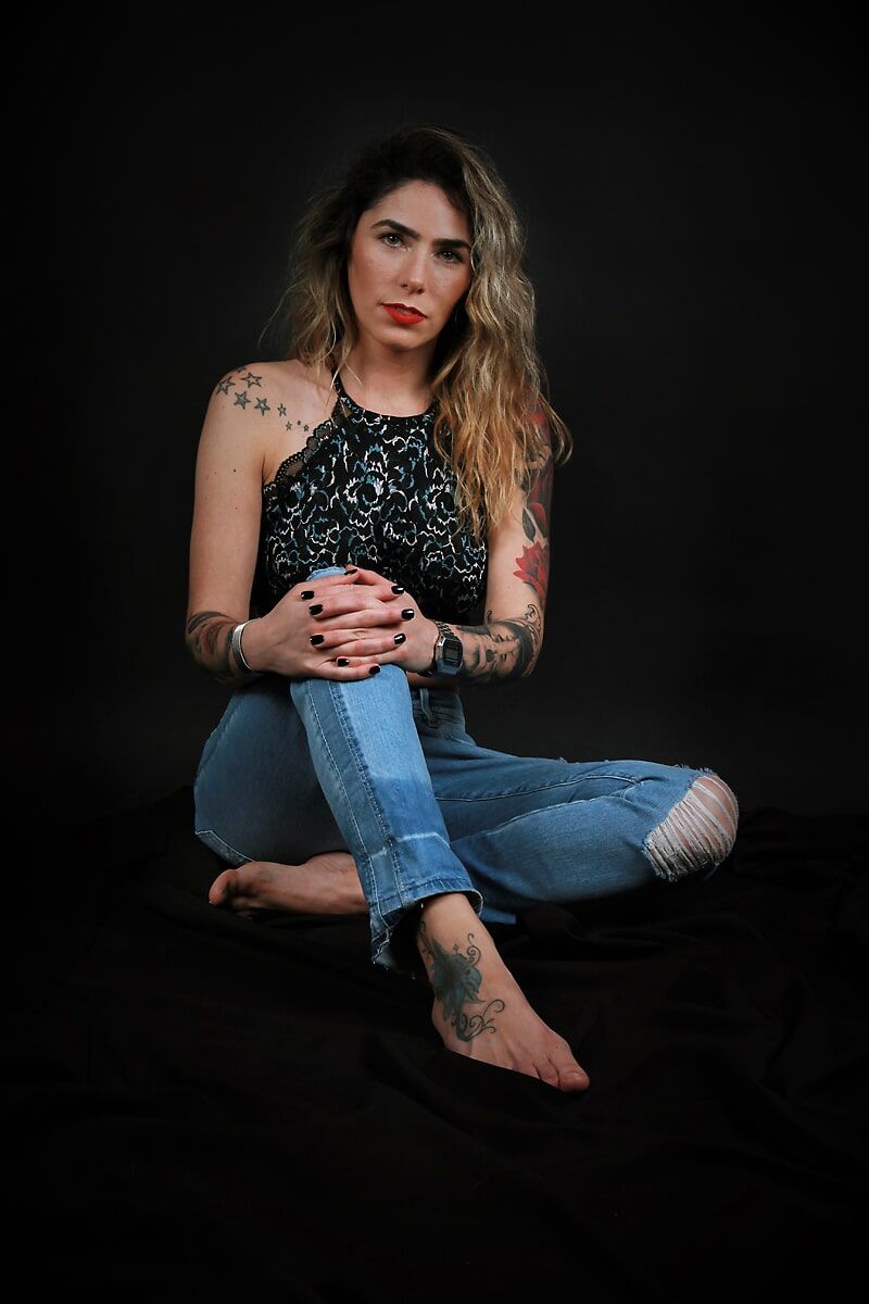 Mulher tatuada, sentada sobre uma superfície preta, usando blusa de alças e calça jeans rasgada, olhando para a câmera.