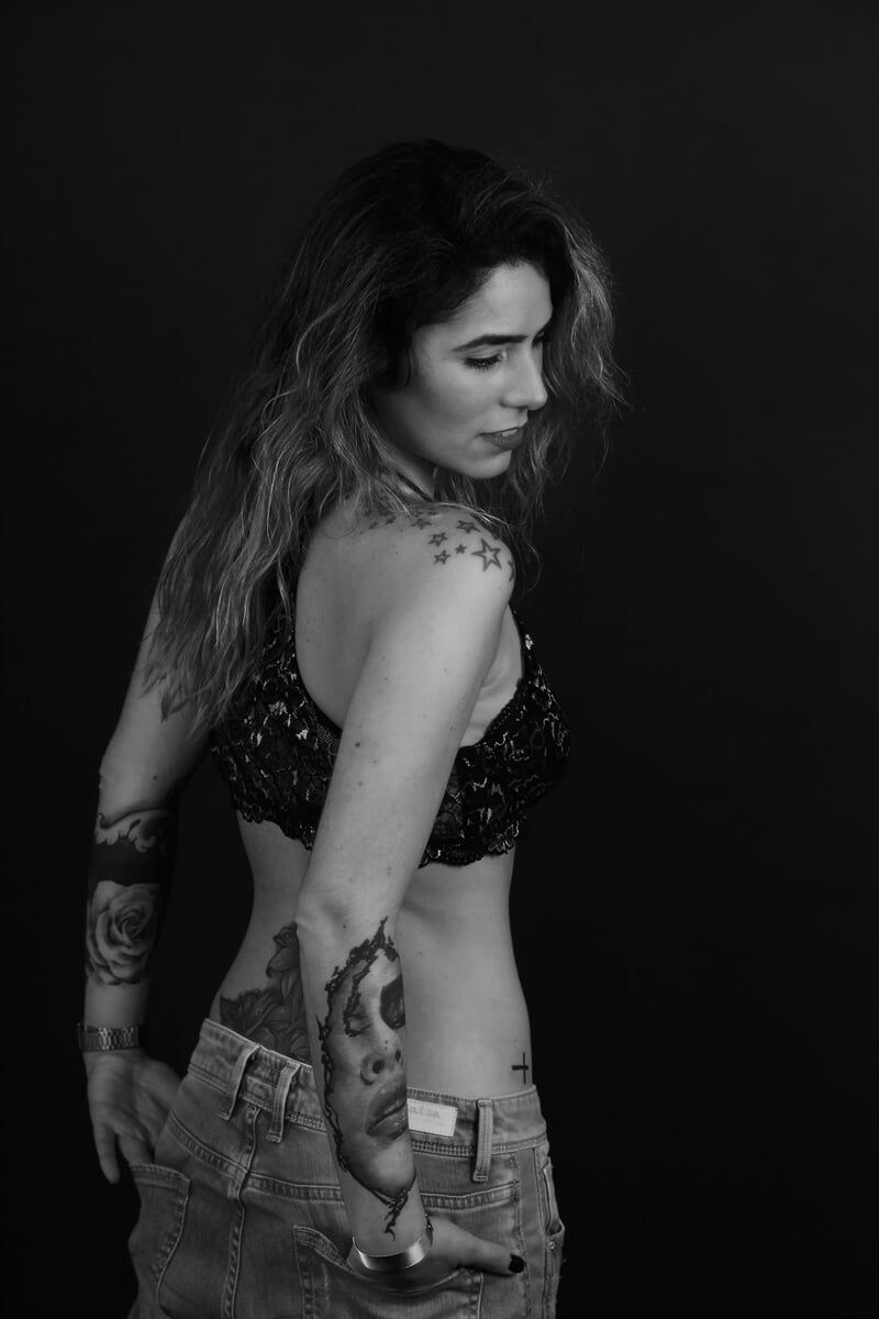 Mulher com tatuagens no braço e nas costas, usando top curto e calça jeans, olhando por cima do ombro.