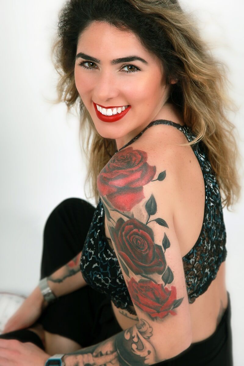 Mulher com tatuagem de rosas vermelhas no ombro sorri, vestindo blusa de miçangas e pulseiras.