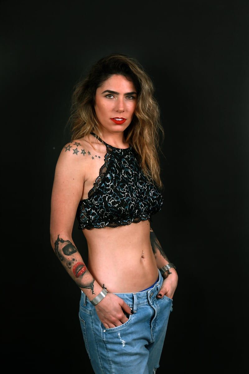 Mulher com tatuagens, mãos nos bolsos, vestindo um top curto e calça jeans, contra um fundo preto.