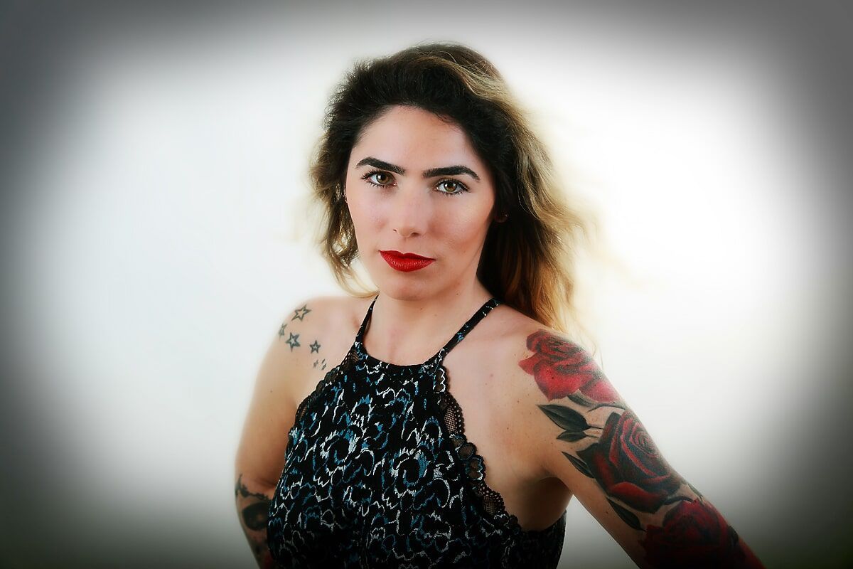 Mulher com batom vermelho e tatuagens no braço, usando um top frente única, posando em frente a um fundo branco.
