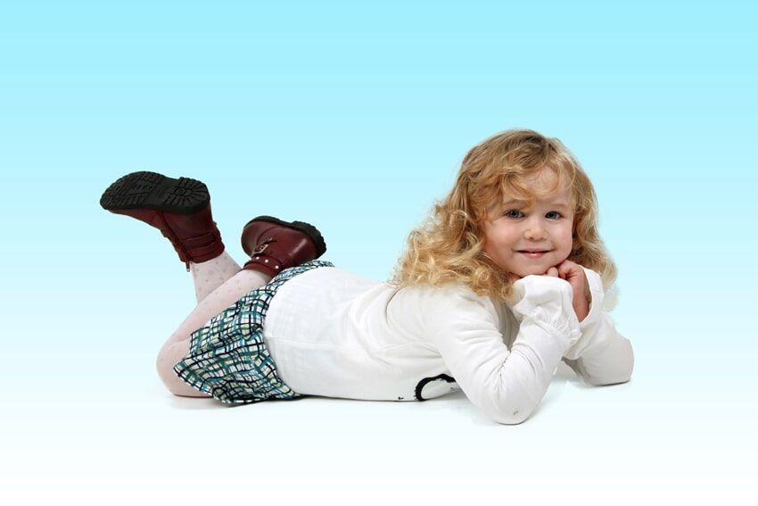 Menina loira de blusa branca e saia xadrez, deitada e sorrindo. Fundo com degradê azul.