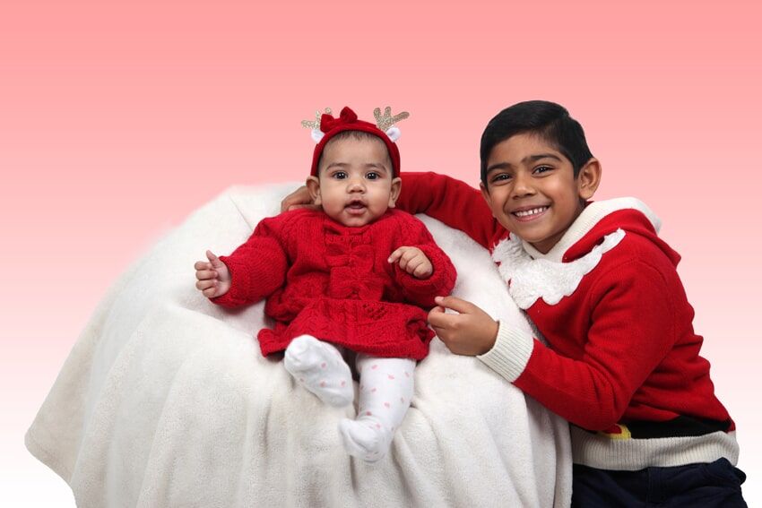 Menino de suéter vermelho sorri ao lado de bebê com chifres de rena e roupa vermelha, ambos em uma almofada branca.