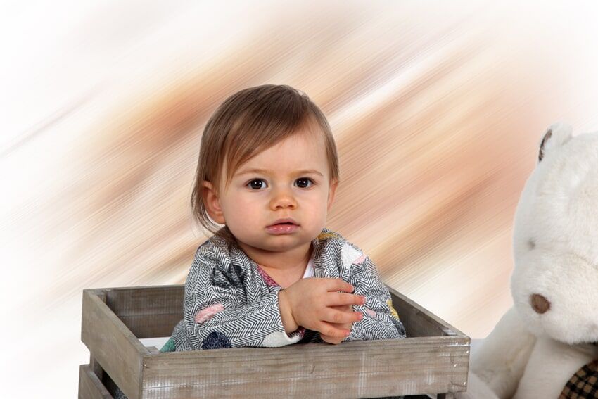 Bebê em uma caixa de madeira com um ursinho de pelúcia, olhando para cima com uma expressão neutra, fundo bege desfocado.