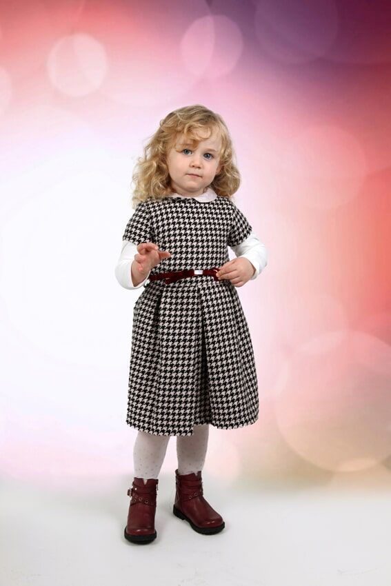 Menina com vestido estampado e botas, em pé contra um fundo colorido com efeito bokeh.