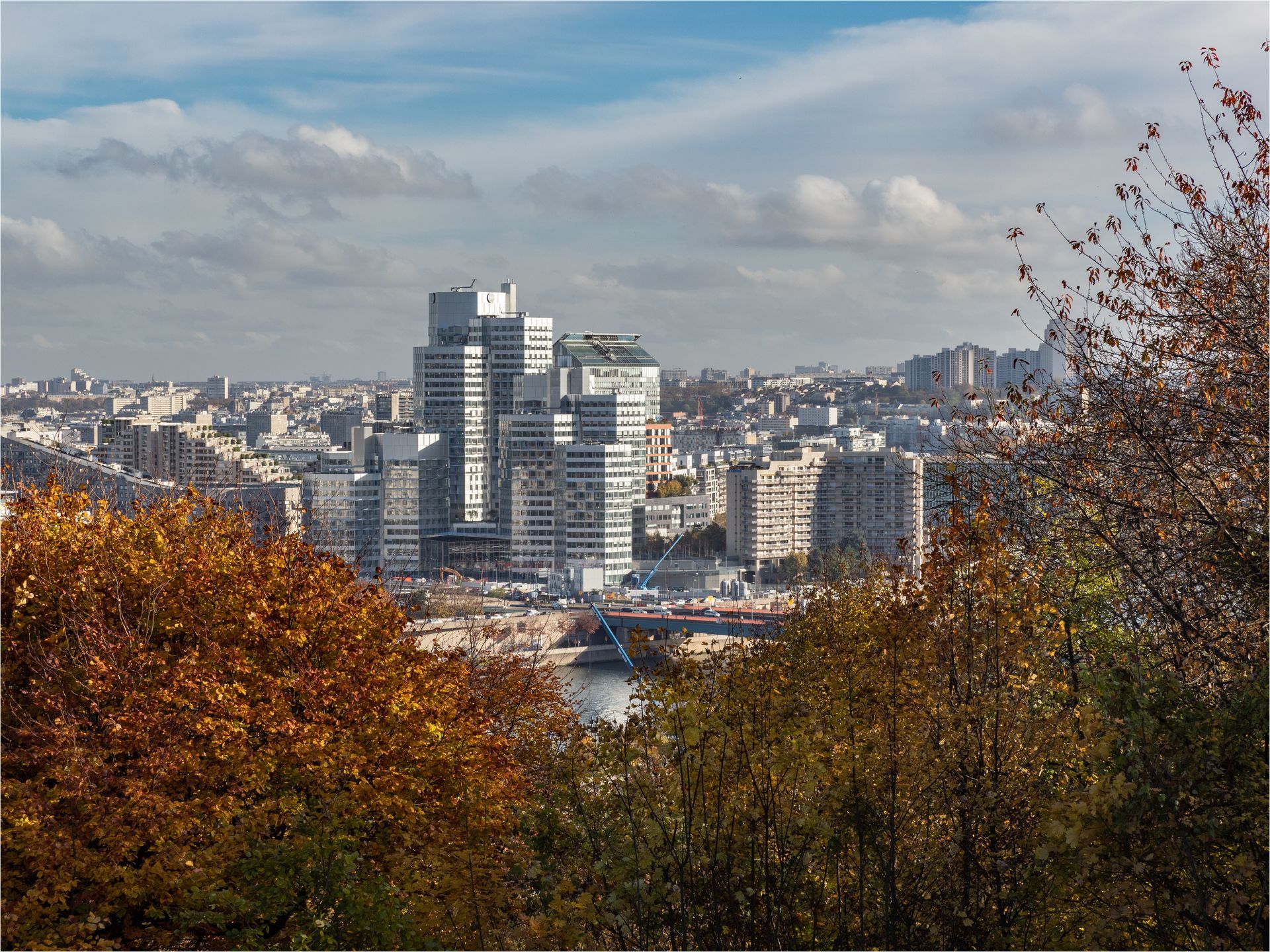 Horizon de la ville avec un groupe de bâtiments modernes à plusieurs niveaux au centre, vu à travers les arbres d'automne sous un ciel nuageux.