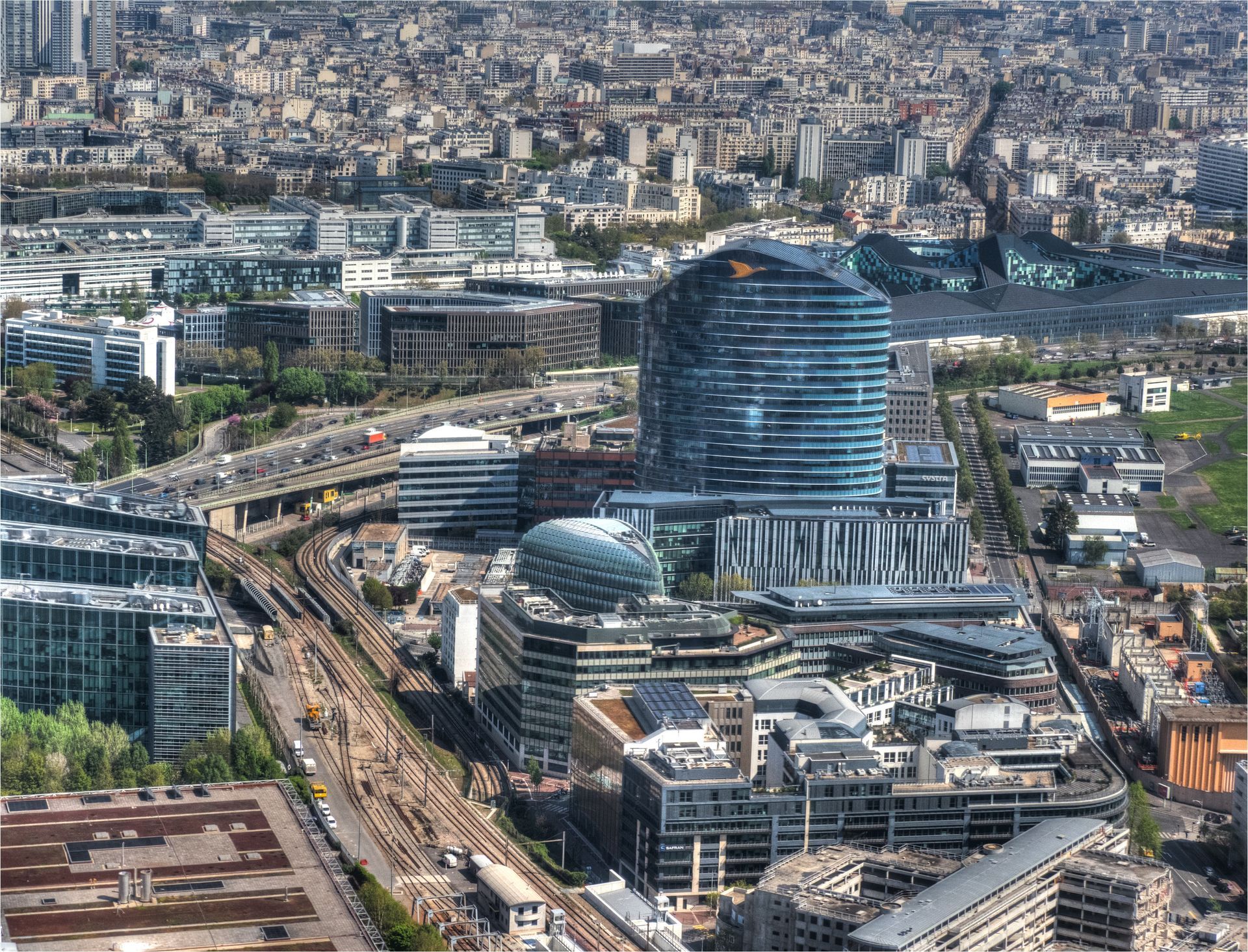Paysage urbain avec des bâtiments modernes, dont un grand gratte-ciel recouvert de verre bleu, et des voies ferrées, Paris.