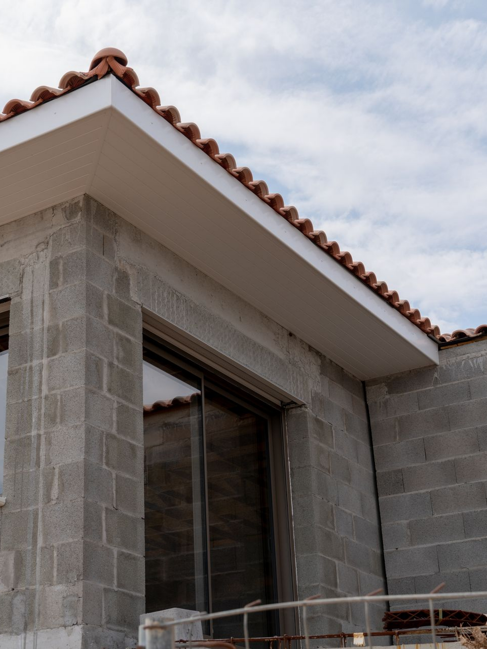 Bâtiment en parpaings gris avec garnitures blanches, tuiles en terre cuite et porte vitrée, contre un ciel nuageux.