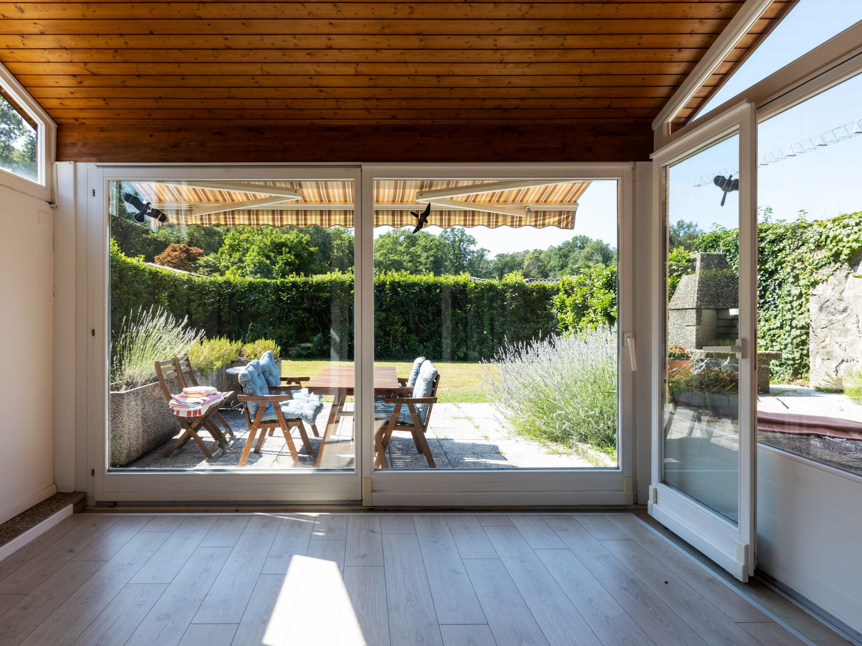 Véranda avec grandes fenêtres donnant sur un jardin, table avec chaises visibles, plafond en bois, porte ouverte.