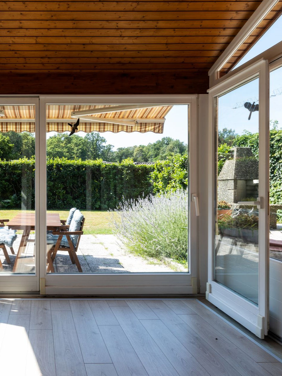 Pièce lumineuse avec portes vitrées donnant sur un jardin. On y découvre un parquet, un plafond marron et du mobilier d'extérieur.