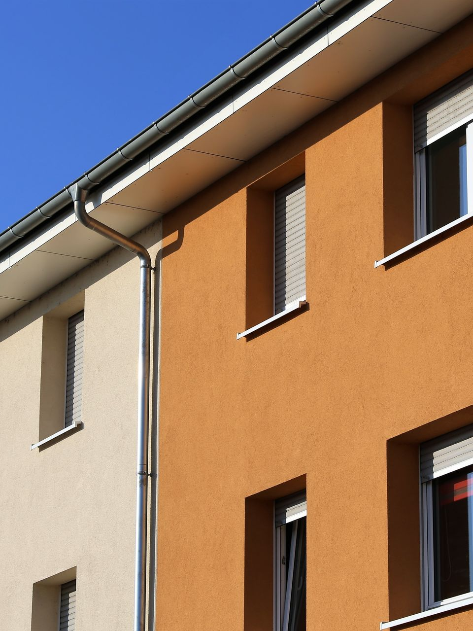 Bâtiments de deux étages aux façades en stuc beige et orange. Fenêtres aux volets blancs et ciel bleu.