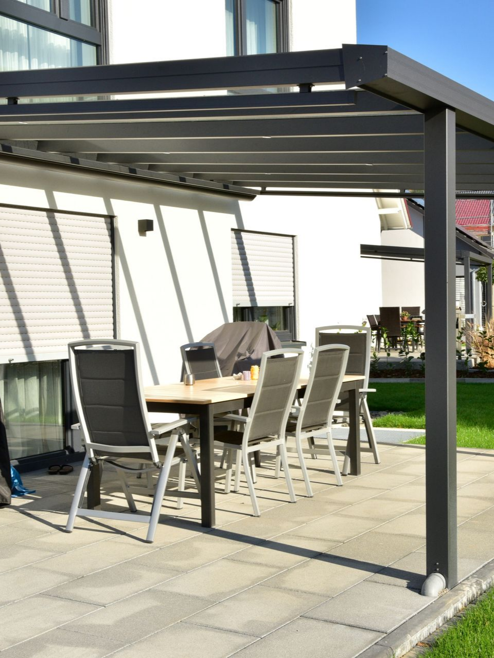 Espace repas extérieur sous une pergola grise, avec table et chaises, sur une terrasse ensoleillée.