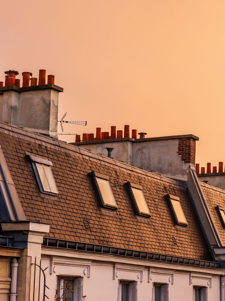 Rangée de bâtiments aux toits de tuiles brunes et aux cheminées contre un ciel couchant.