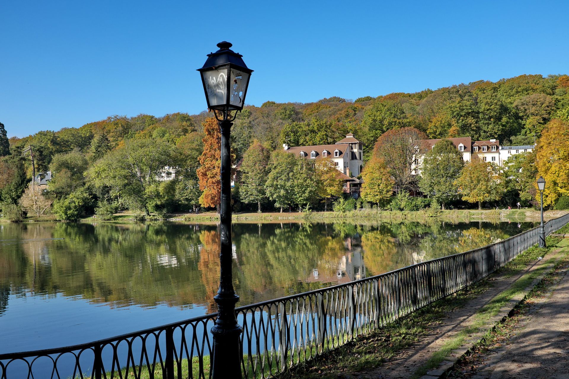 Un lac avec des feuilles d'automne, un lampadaire et un bâtiment au loin. Ciel bleu et reflets dans l'eau.