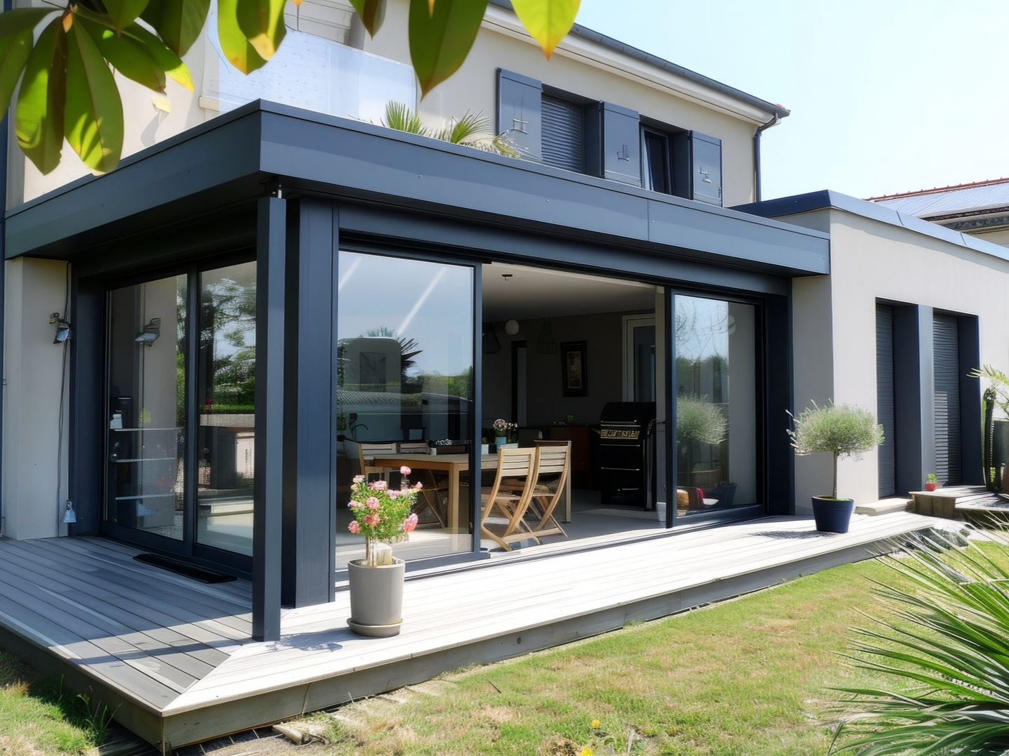 Maison moderne avec une extension grise et de grandes portes coulissantes en verre ouvrant sur une terrasse en bois, journée ensoleillée.