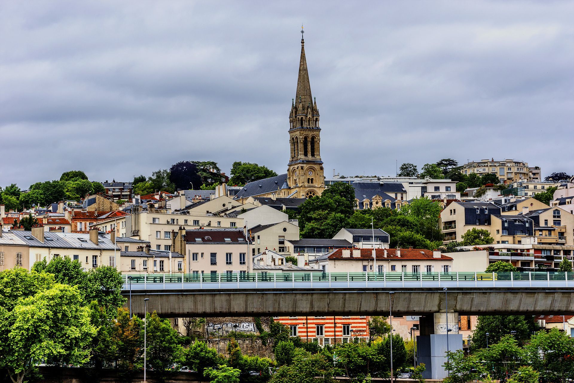 Paysage urbain avec un haut clocher d'église, un ciel nuageux et un pont au premier plan.