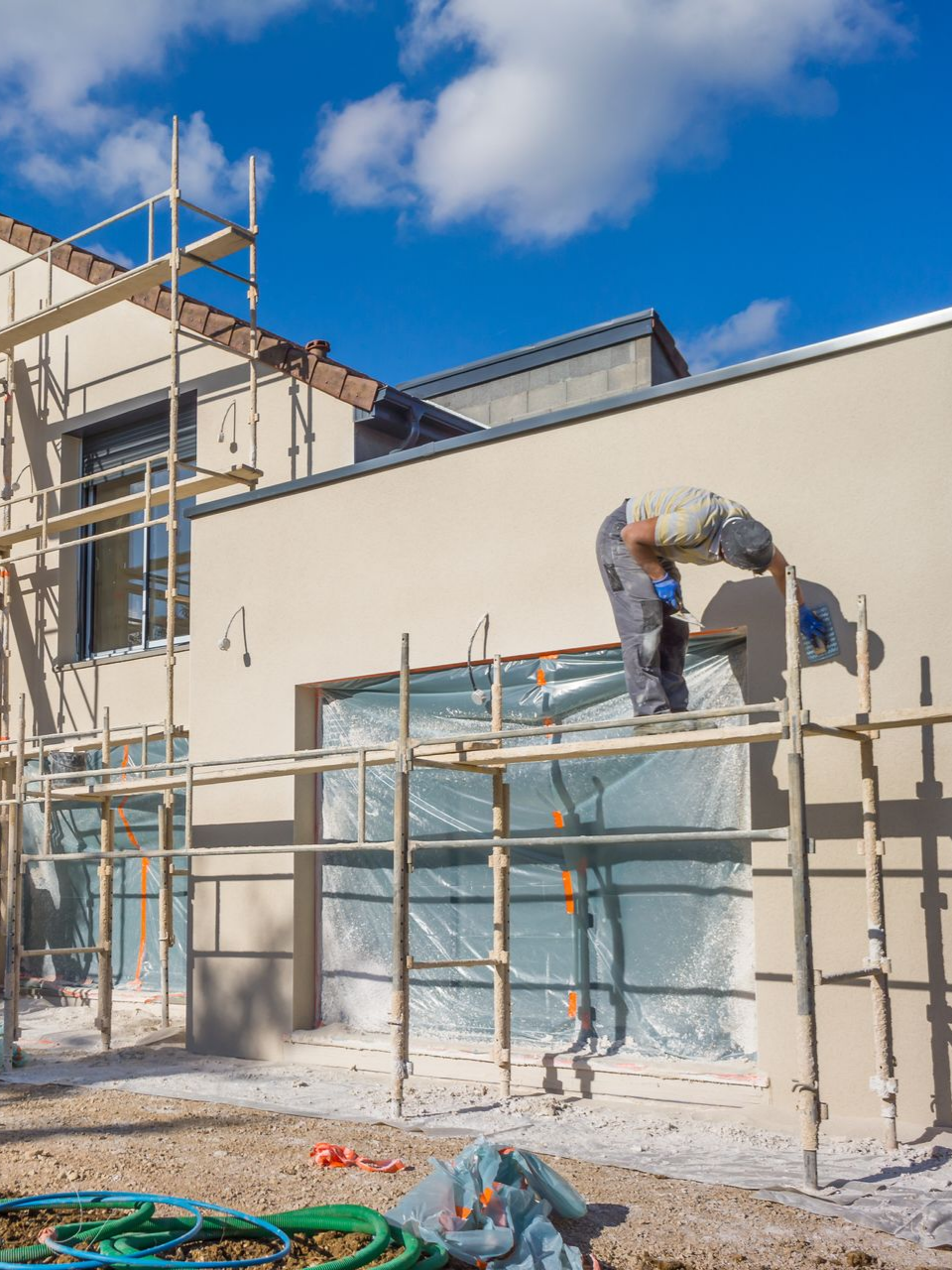 Ouvrier du bâtiment sur un échafaudage en train de plâtrer une façade de bâtiment beige sous un ciel bleu.