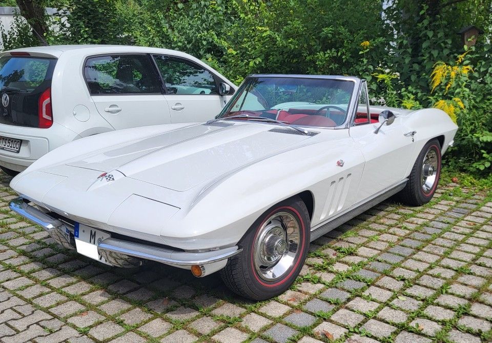 Ein weißes Corvette-Cabriolet mit roter Innenausstattung parkt auf einer gepflasterten Fläche neben einem weißen Auto.