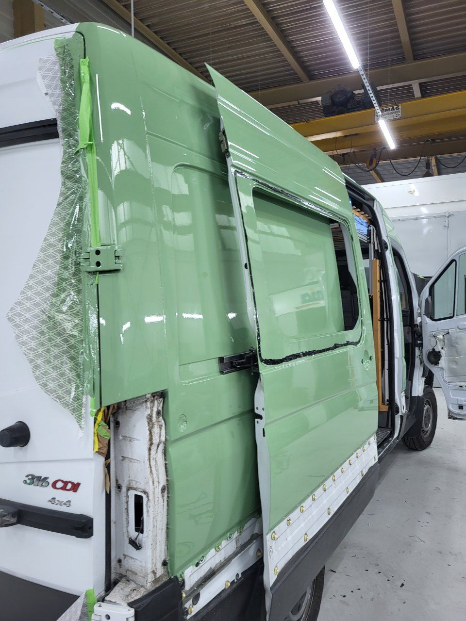 In einer Werkstatt wird an einem grün lackierten Kastenwagen-Karosserieteil gearbeitet, Fensterausschnitt, weiß und grün.