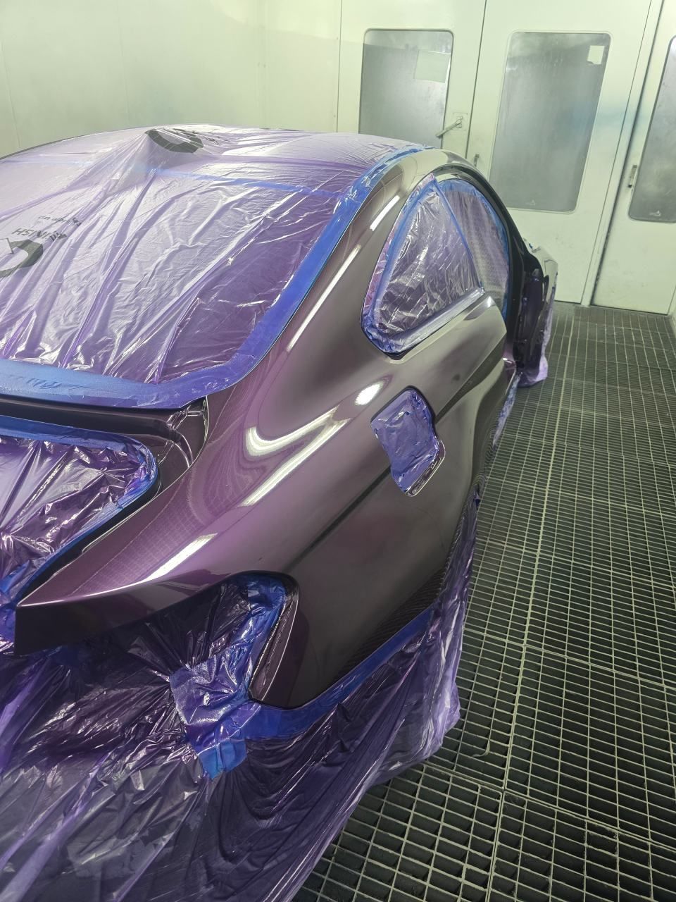 Ein Auto in einer lila Lackierkabine, teilweise mit Plastikfolie abgedeckt, mitten im Lackiervorgang.