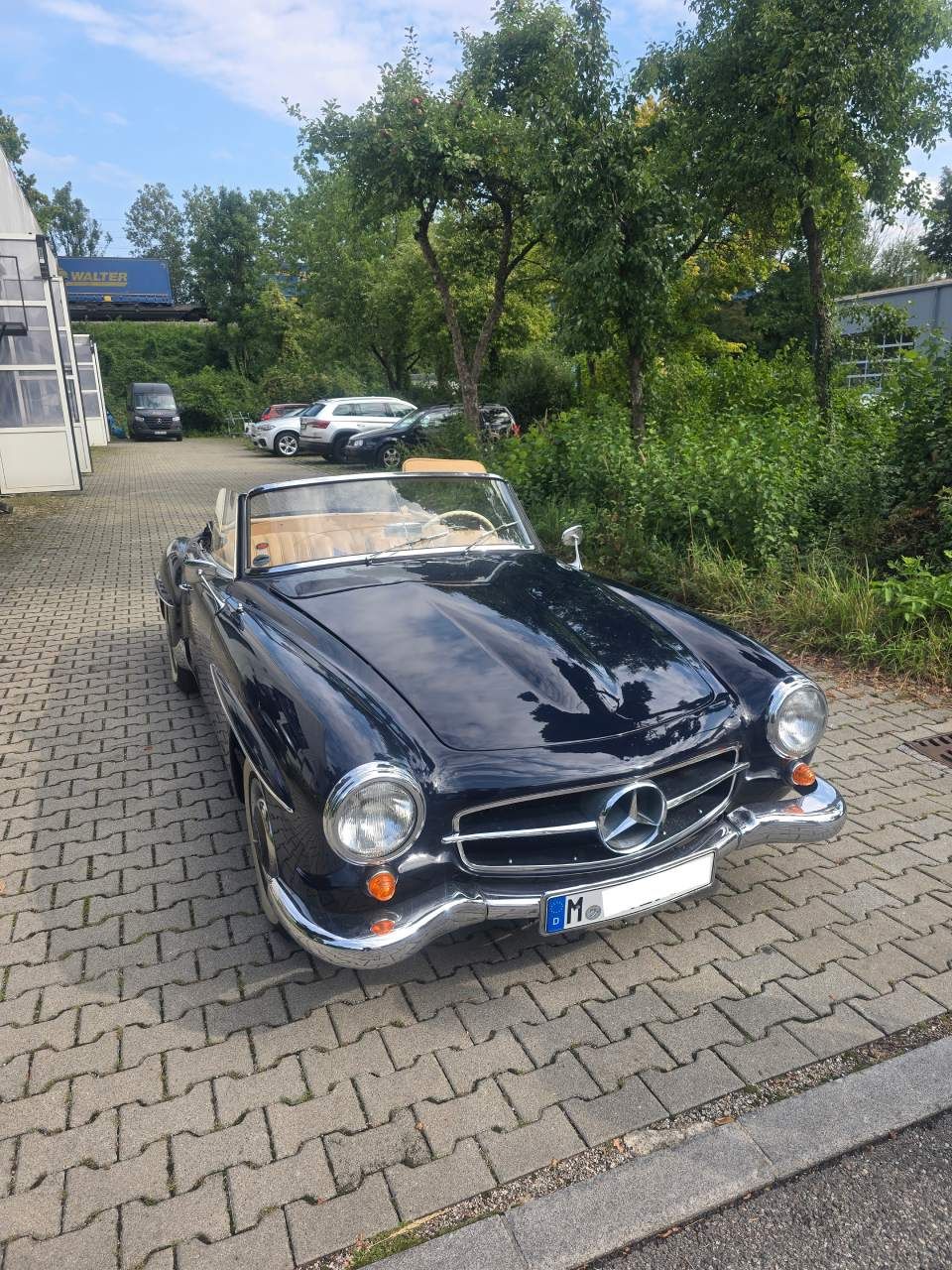 Ein dunkelblaues Oldtimer-Cabriolet von Mercedes-Benz parkt auf einem gepflasterten Platz, im Hintergrund ist Grün zu sehen.