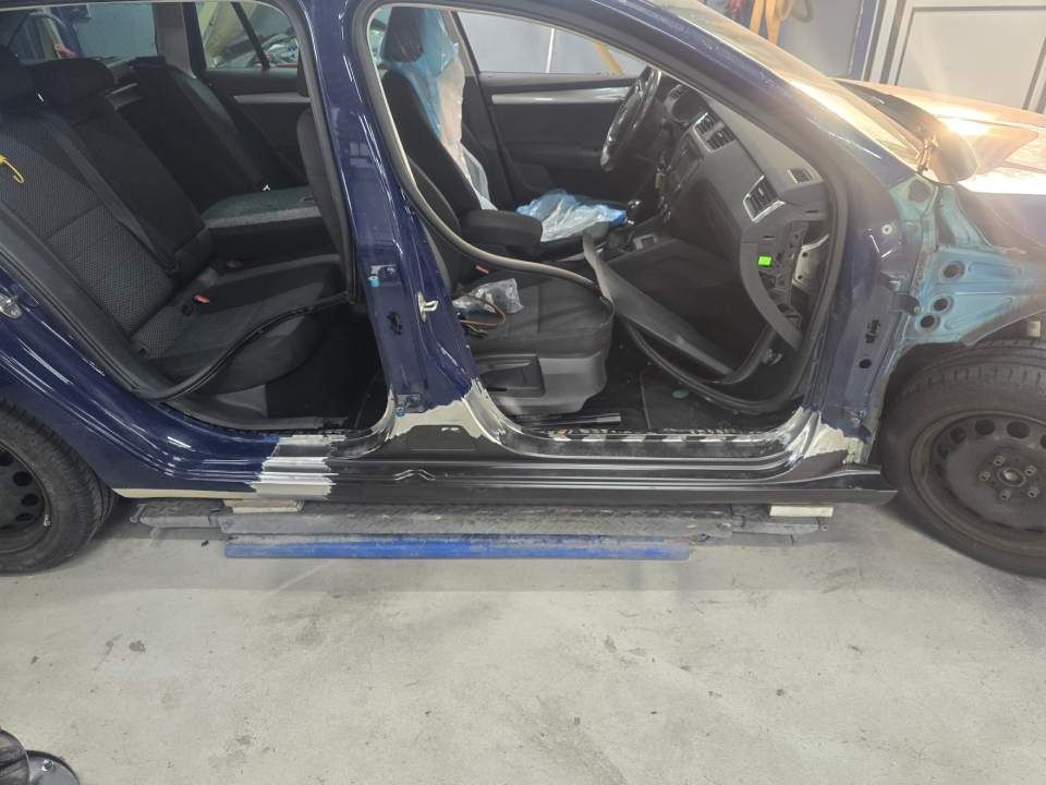 Blaues Auto mit beschädigter Seite, wird in einer Werkstatt repariert. Tür ausgebaut, Innenraum sichtbar.