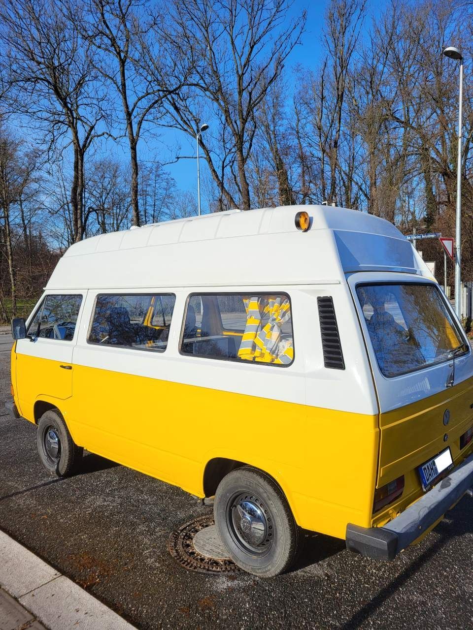 Ein gelb-weißer VW-Transporter parkt in einer Straße mit kahlen Bäumen und klarem blauen Himmel.