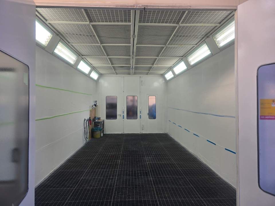 Innenansicht einer leeren, weißen Lackierkabine mit Gitterboden, Deckenbeleuchtung und Doppeltüren.