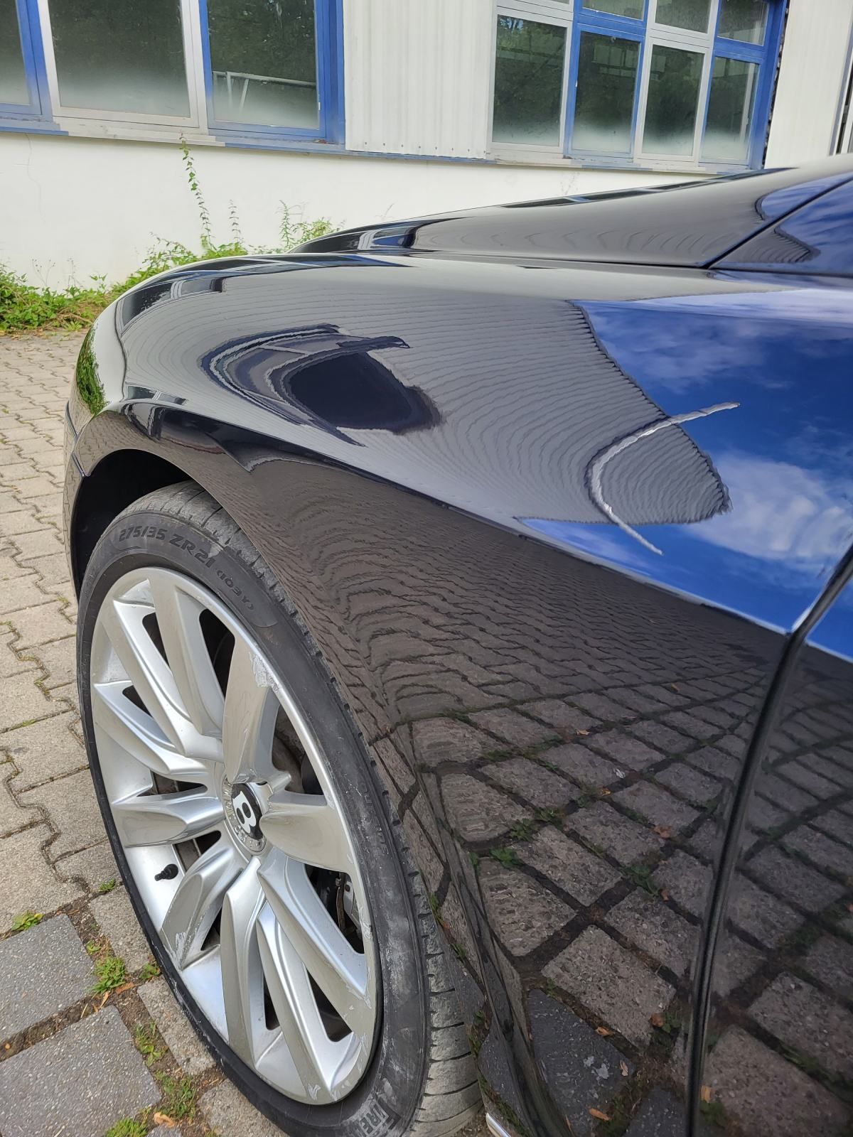 Schwarzer Kotflügel mit Reifen und silberner Felge, der Gebäude und Himmel spiegelt.