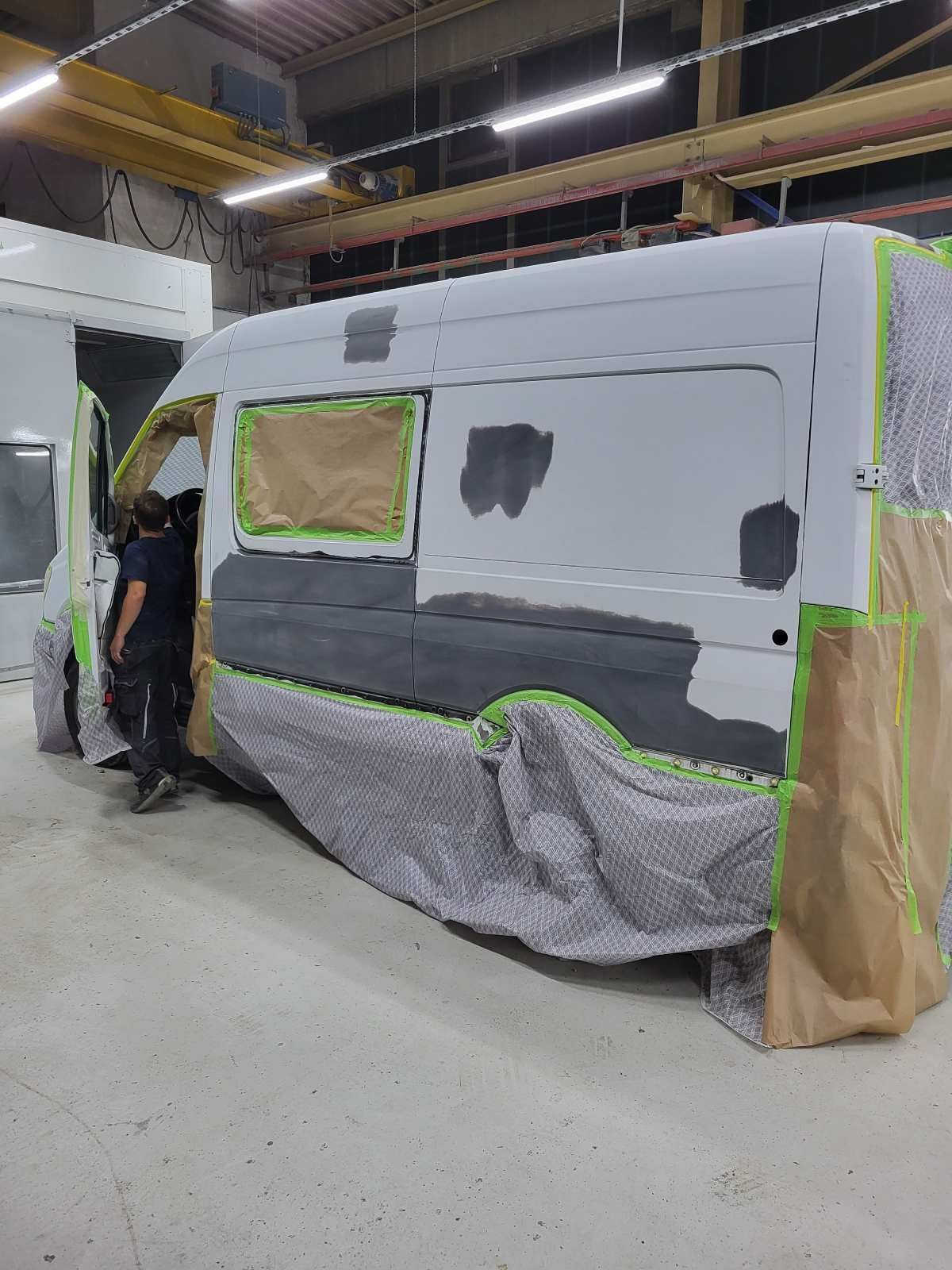 Ein weißer Lieferwagen in einer Werkstatt, teilweise mit Schutzfolie und Klebeband verhüllt. Eine Person steht daneben.