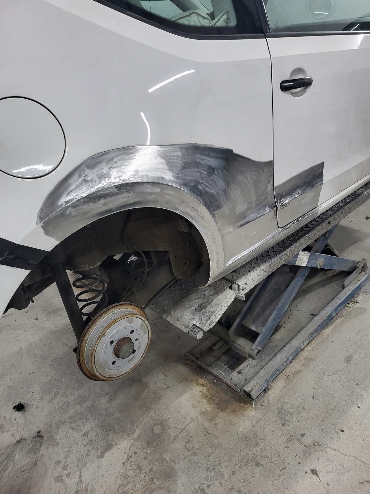 Weißes Auto mit Karosseriereparaturen am hinteren Kotflügel, auf einem Wagenheber abgestützt.