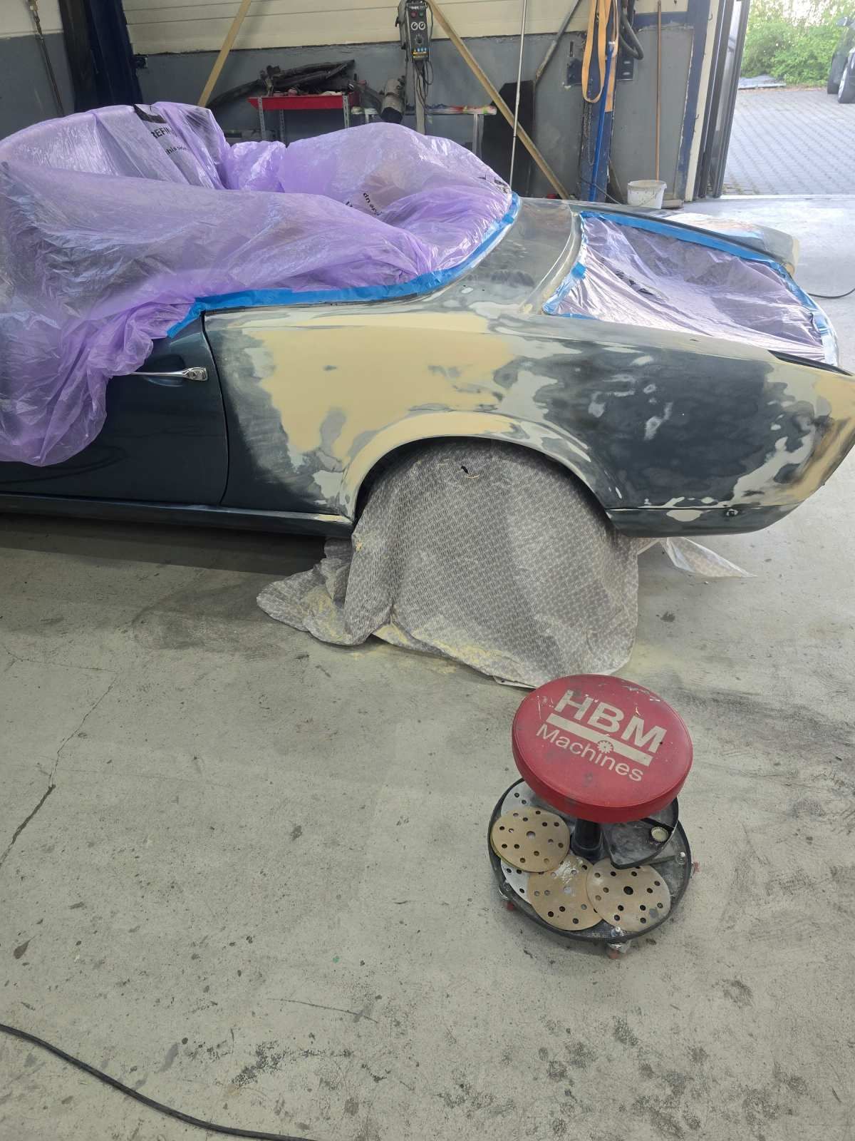 In einer Garage wird an einem Auto gearbeitet, der Kotflügel ist geschliffen und grundiert. Lila Abdeckung, roter Hocker, Betonboden.