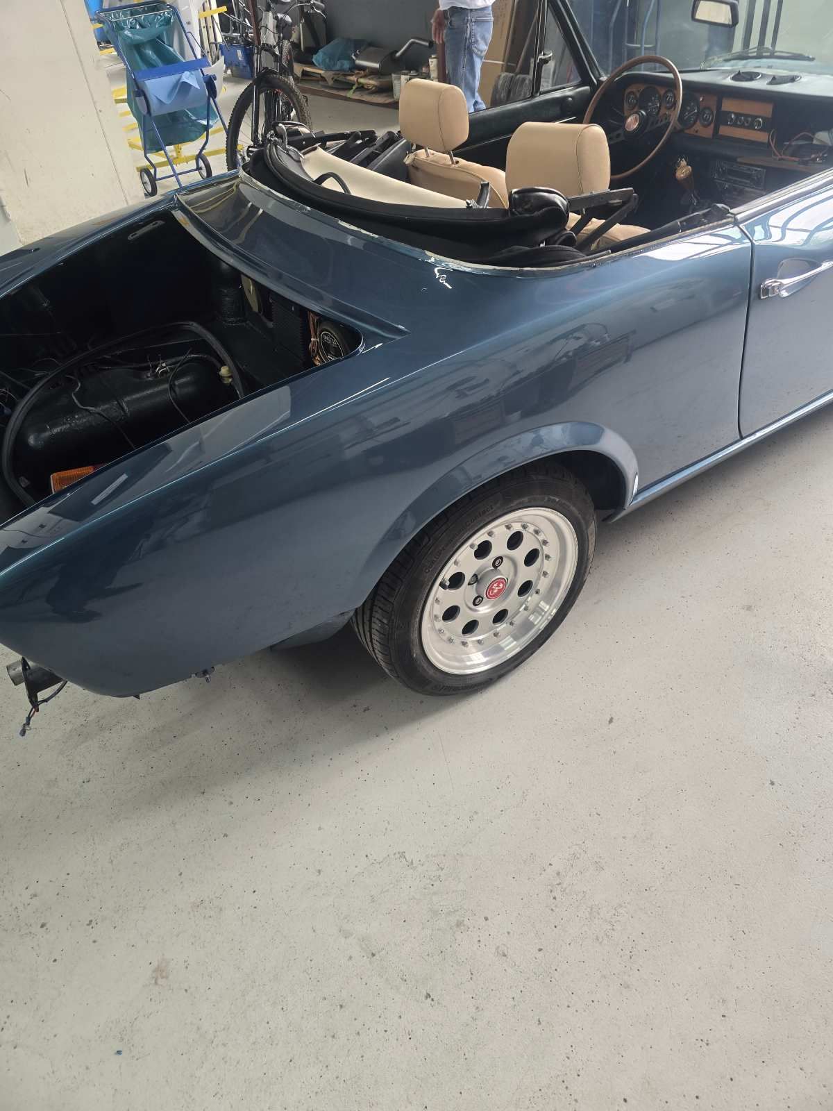 Blaues Cabriolet mit geöffneter Motorhaube, die Motorteile freigibt. Silberne Felgen, beige Sitze. Innenaufnahme.