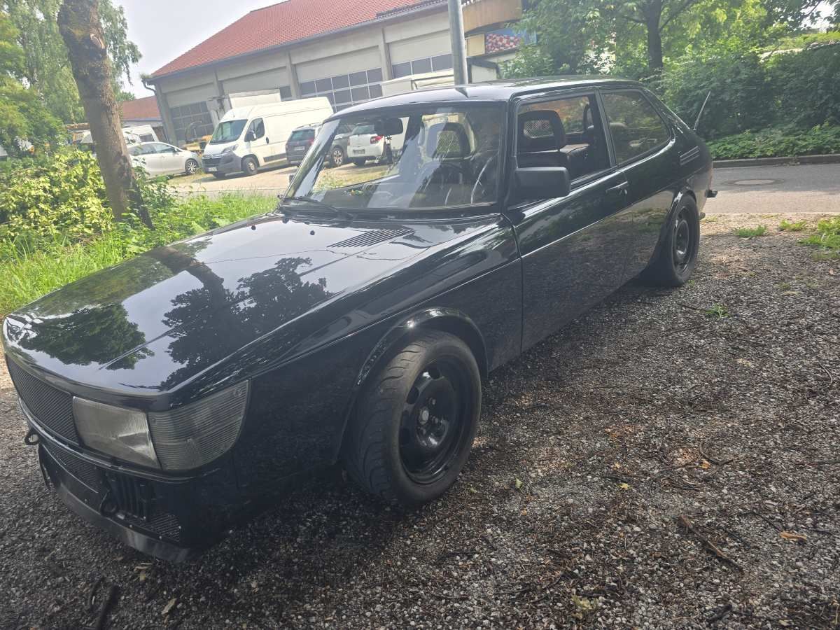 Ein schwarzes Saab 900 Coupé parkt auf einer Schotterauffahrt. Im Hintergrund sind eine Fabrik, Bäume und ein Lieferwagen zu sehen.
